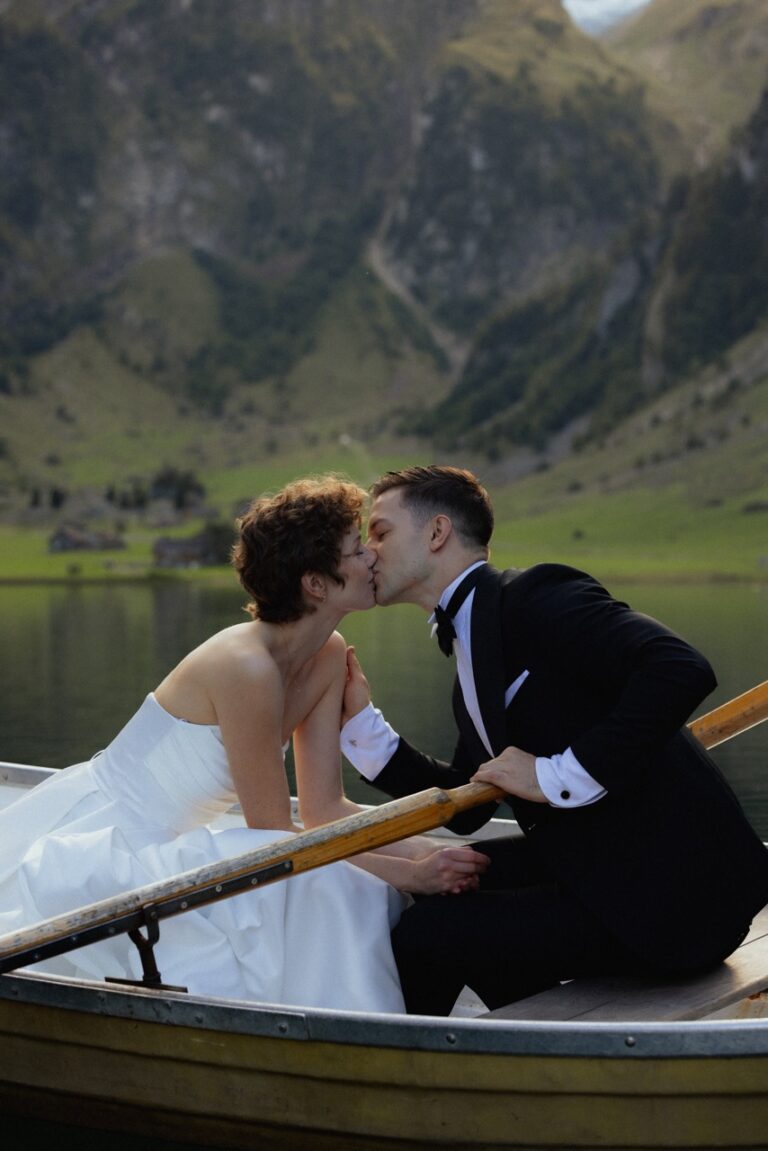 groom and bride sharing a kiss on a wooden paddle boat on the lake with mountain behind them